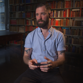 A man seated with a beard with a book case in the background
